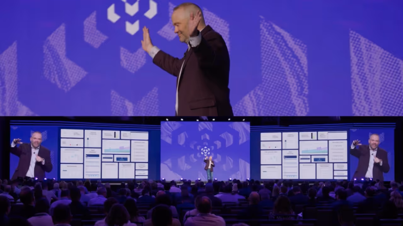 Speaker on stage presenting with multiple large screens displaying charts and data to a seated audience in a conference setting.
