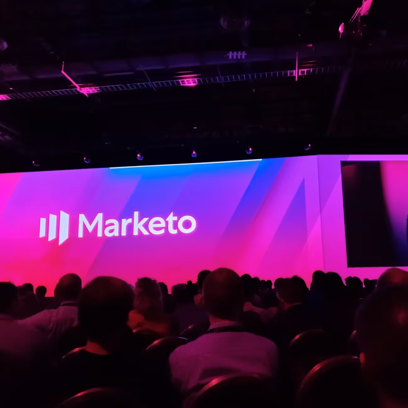 Audience seated in a dark room facing a large screen displaying the Marketo logo with pink and purple lighting.