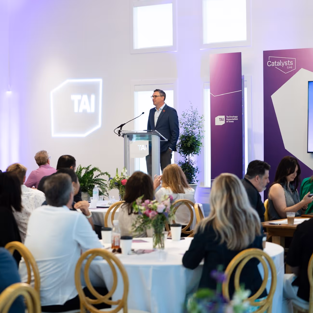 Man in suit speaking at a podium during a Technology Association of Iowa event with seated audience watching.
