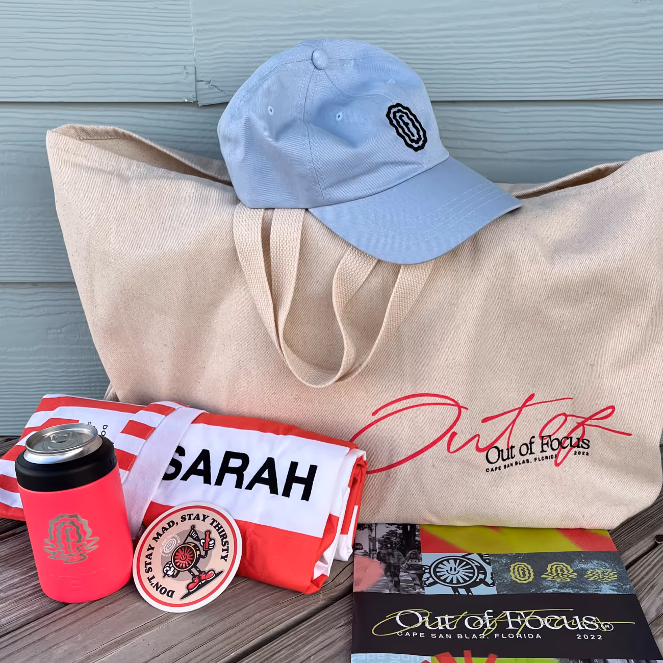 Beige tote bag with red and black 'Out of Focus' text, light blue cap on top, red and white folded cloth, red can cooler, round sticker, and colorful brochure on wooden surface.