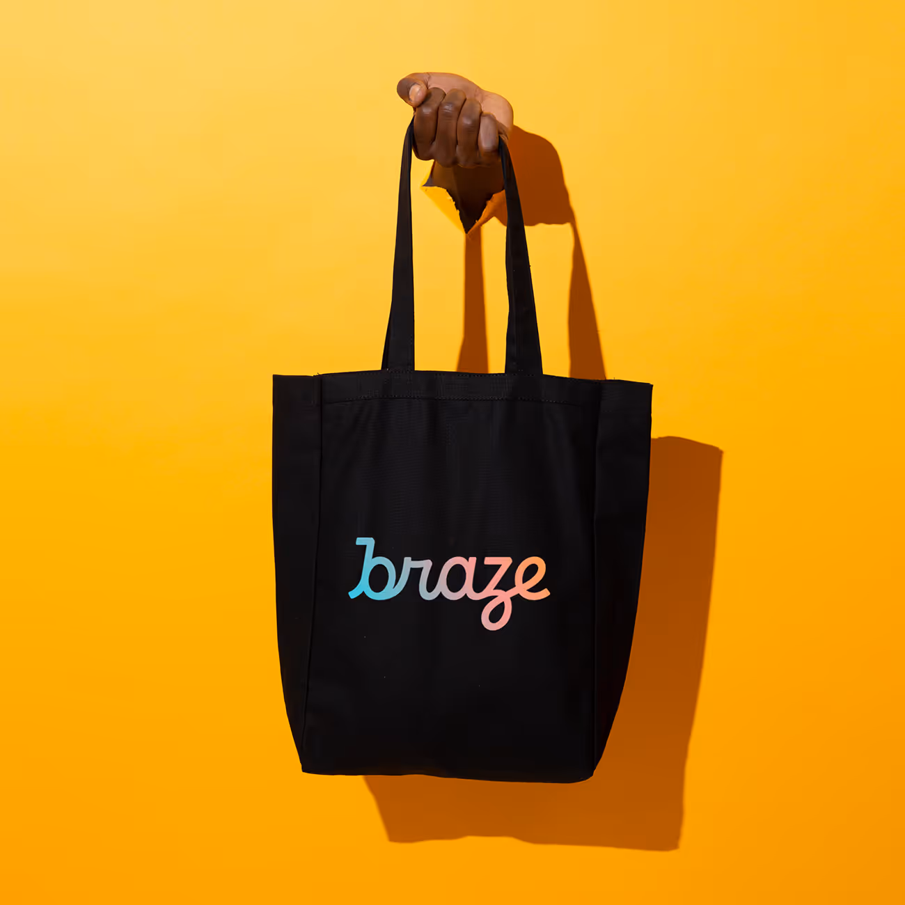 Hand holding a black tote bag with the word 'braze' in blue and pink lettering on a yellow background.