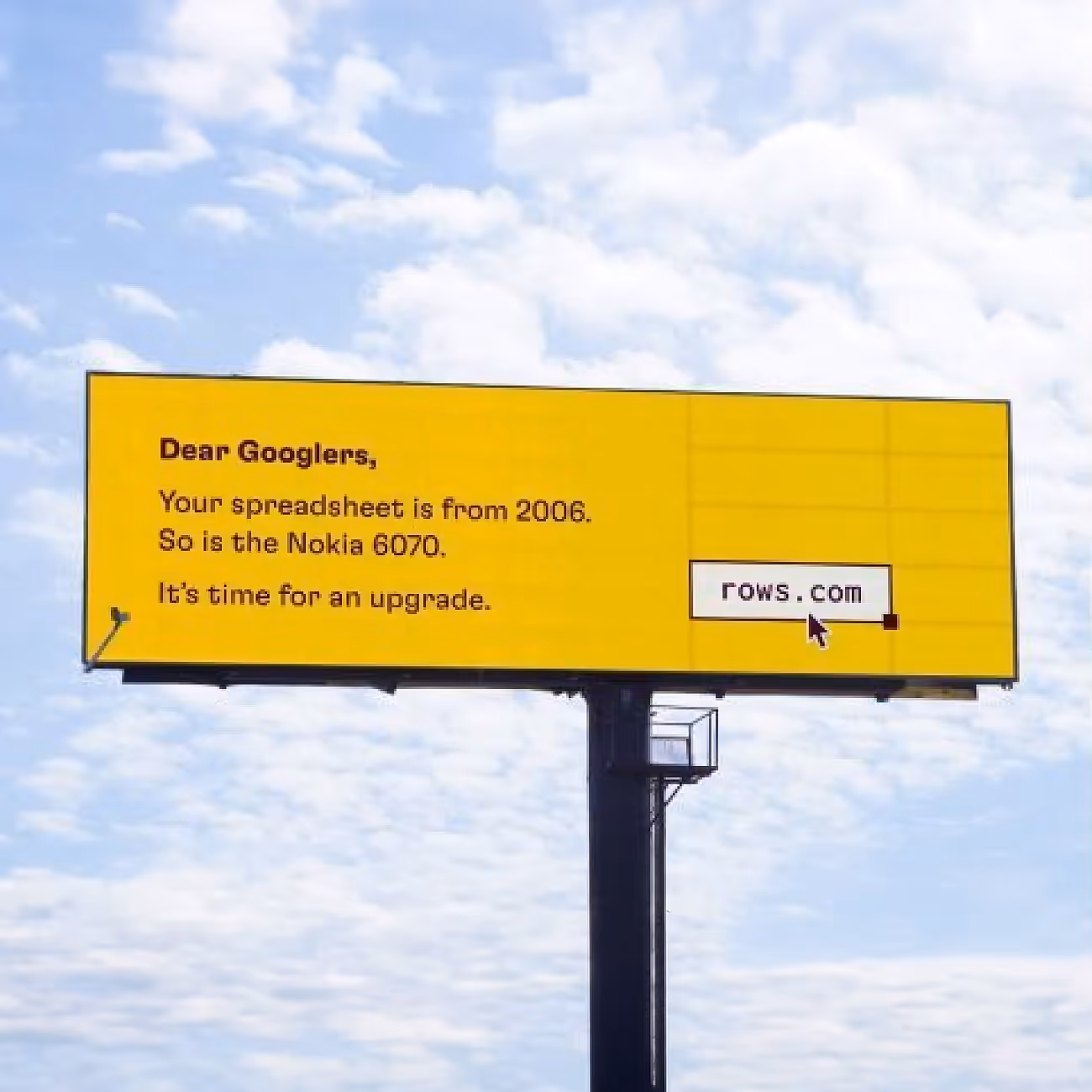 Yellow billboard against a blue sky with text urging Google users to upgrade their spreadsheet, referencing its 2006 origin and comparing it to a Nokia 6070, with rows.com displayed.